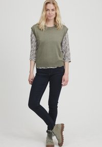 Olivenfarvet ærmeløs top over en blomstermønstret langærmet skjorte, parret med mørke jeans og grå-brune snørebåndsstøvler.