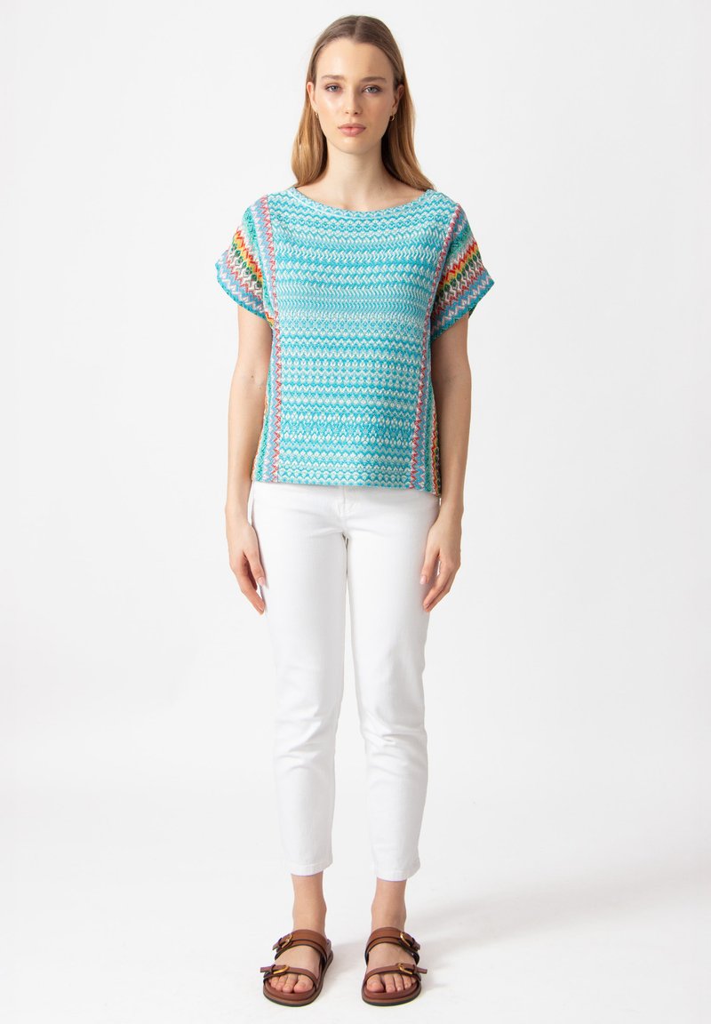 Top azzurro in maglia a maniche corte, con un motivo a zig zag e accenti colorati lungo i lati, abbinato a pantaloni bianchi e sandali marroni.