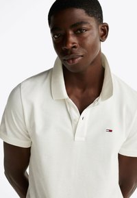 Camiseta polo blanca con cuello clásico, mangas cortas y un pequeño logo bordado en rojo, blanco y azul en el pecho. Tejido texturizado.