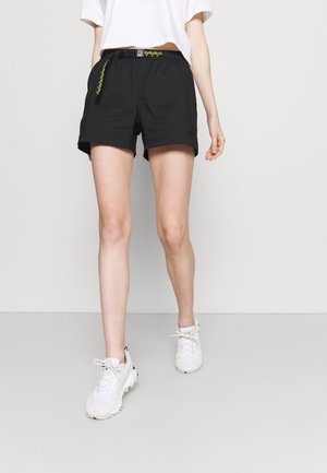 Person iført sorte shorts med gul akcent på bæltet, hvid cropped t-shirt og hvide atletiske sneakers på en ensfarvet baggrund.