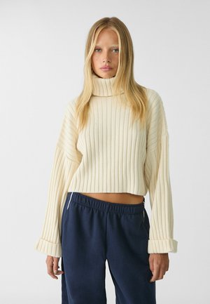 Light beige, ribbed turtleneck sweater with a cropped fit and wide sleeves, paired with navy relaxed-fit pants. Textured fabric detail visible.