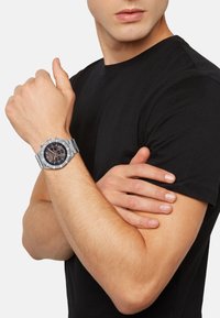 Montre chronographe argentée sur un poignet, arborant un cadran noir, des marqueurs lumineux et un bracelet en acier inoxydable. Portée par-dessus une chemise noire.