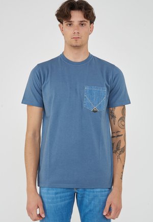 Giovane uomo con capelli corti castani che indossa una maglietta blu con una tasca in denim e jeans blu, in piedi davanti a uno sfondo bianco.