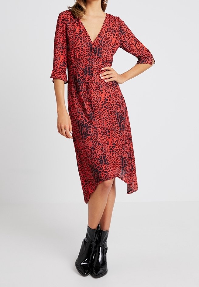 Robe rouge à imprimé léopard avec un col en V, des manches trois-quarts et un ourlet asymétrique. Portée avec des bottines noires brillantes.