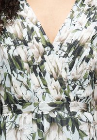 Φλοράλ φόρεμα τύπου wrap με ανοιχτό γαλάζιο φόντο, διακοσμημένο με λευκά και πράσινα μοτίβα μαγνόλιας και χαρακτηριστικό σχήμα που σφίγγει στη μέση. Μαλακό, υφασμάτινο με υφή.