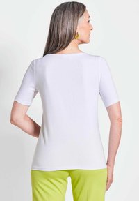 Weißes kurzärmliges Hemd mit Rundhalsausschnitt, aus glattem Stoff, getragen mit grünen Hosen, zeigt ein einfaches, sauberes Design.