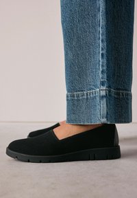 Jalat memakai sepatu slip-on hitam yang memiliki tekstur, dipadukan dengan jeans denim biru dengan hem lebar, berdiri di atas lantai abu-abu.