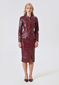 Veste en simili cuir bordeaux avec fermeture éclair à l'avant et col montant, assortie d'une jupe avec une fente à l'avant. Des chaussures à talons hauts complètent le look.