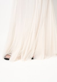 Lichtbeige doorschijnende stoffen rok met vloeiende lagen, die een zwarte hoge hak onthult, versierd met een strass-detail op de hak.