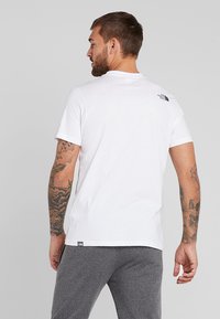 Valge lühikeste varrukatega t-särk, mis on valmistatud puuvillast, väikese musta logoga selja ülaservas. Modell kannab halli pükse ja tal on tätoveeringud.