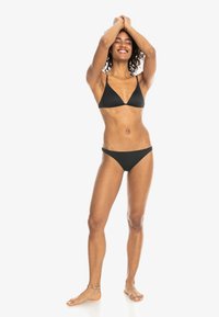 Black bikini set with a triangular top and low-rise bottom, made from smooth material. Model stands barefoot, arms raised, smiling.