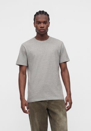 Ung mand med fletninger iført en ensfarvet lysegrå kortærmet t-shirt og olivengrønne bukser, stående mod en hvid baggrund.