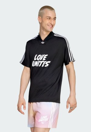 Lächelnde Person mit schwarzem Adidas-Shirt und Shorts mit pastellfarbenem Farbverlauf, beide mit der Aufschrift „LOVE UNITES“ und Adidas-Streifen.