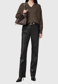 Brązowy sweter z dekoltem w serek, czarne skórzane spodnie i czarne szpilki z ostrym noskiem. Trzyma ciemnoszarną torebkę. Gładka tkanina, dopasowany krój, dopasowana sylwetka.