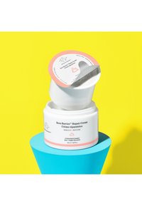 Un pot de crème blanche étiqueté "Crème Réparatrice Bora Barrier" avec un couvercle rose, placé sur un piédestal bleu contre un fond jaune lumineux.