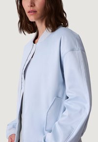 Jasnoniebieska kurtka bomberka z gładkiej tkaniny, z zamkiem błyskawicznym, ściągaczem przy kołnierzu oraz bocznymi kieszeniami, o luźnym kroju i minimalistycznym designie.