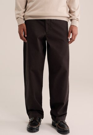 Homme portant un pantalon noir ample, un pull beige et des chaussures à lacets noires, debout sur un fond uni clair.