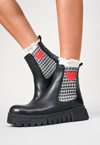Stivali ankle in pelle nera con pannelli in houndstooth elastico che presentano cuori rossi, polsini bianchi svasati e suole in gomma spessa con scanalature.