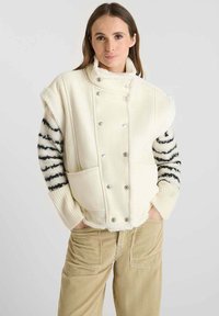 Crème kleurige vest met drukknoopjes, grote voorzakken en nepbontafwerking. Mouwen hebben een zwart-wit gestreept patroon.