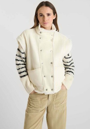 Gilet color crema con bottoni a pressione, grandi tasche frontali e bordo in pelliccia sintetica. Le maniche presentano un motivo a righe bianche e nere.