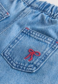 Denim bleu avec une ceinture élastique et une poche, avec des surpiqûres rouges et un petit nœud rouge brodé sur la poche.