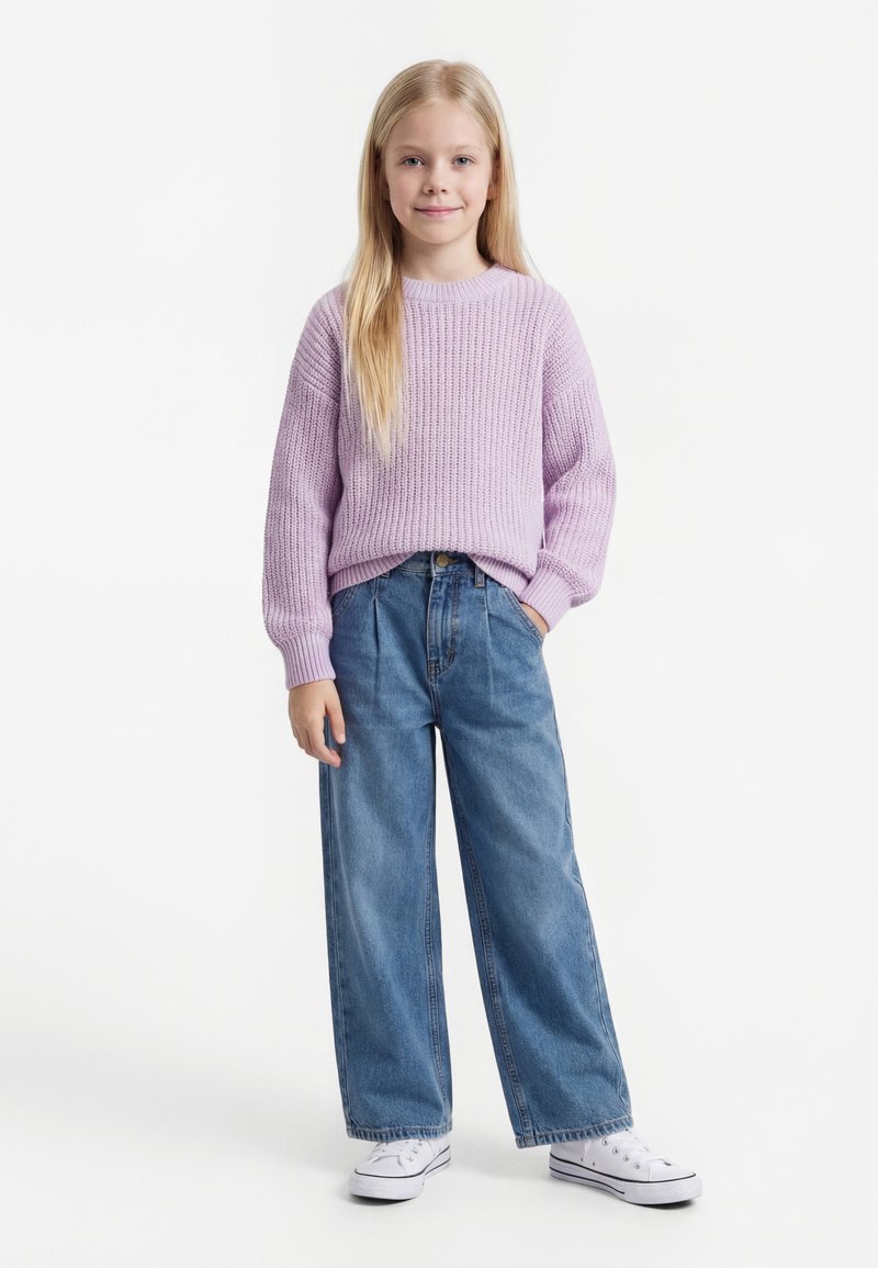 Jeune fille aux longs cheveux blonds portant un pull en laine lilas, un jean large bleu et des baskets blanches, debout avec une main dans la poche.