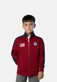 Chaqueta roja con cremallera y mangas en azul marino, que presenta un parche de la bandera estadounidense y un logo redondo en el pecho. Tejido suave con un diseño deportivo.