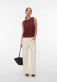 Femme portant un haut sans manches en maille bordeaux, un pantalon large couleur crème, des talons ouverts noirs, tenant un sac à main noir sur un fond blanc.