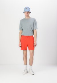 Maglietta grigia a maniche corte, pantaloni corti rossi brillanti con coulisse, cappello a secchiello blu con logo, sneaker bianche e calzini bianchi.
