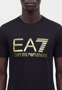 Άνδρας που φοράει μαύρο Τ-shirt με μεγάλα χρυσά γράμματα "EA7 Emporio Armani" τυπωμένα στο στήθος.