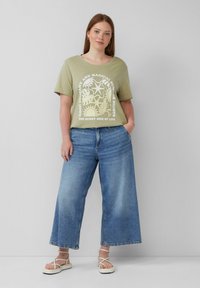 Olivengrünes T-Shirt mit grafischem Druck, kombiniert mit weit geschnittenen blauen Jeans. Modell trägt leichte Sandalen und steht vor einem neutralen Hintergrund.