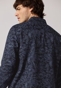 Námořnická modrá košile s texturovaným vzorem napodobujícím listoví. Disponuje klasickým límcem a detailem na zadním dílu, vyrobena z lehkého materiálu.