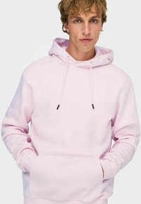 Felpa con cappuccio rosa chiaro con coulisse, tasca frontale e polsini elasticizzati. Texture liscia con design minimalista e dettagli neri.