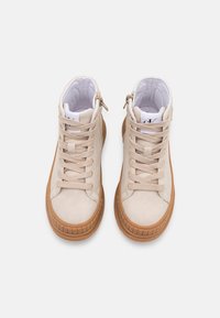 Para beżowych sneakersów za kostkę z gumowymi podeszwami i beżowymi sznurówkami, widziana z góry na jednolitym białym tle.