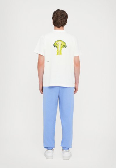 Weißes Baumwoll-T-Shirt mit einem grünen Brokkoli-Design auf dem Rücken, kombiniert mit hellblauen Sweatpants und weißen Sneakern.