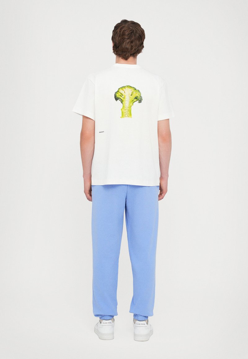 T-shirt blanche en coton avec un graphique de brocoli vert au dos, associé à un pantalon de survêtement bleu clair et des baskets blanches.