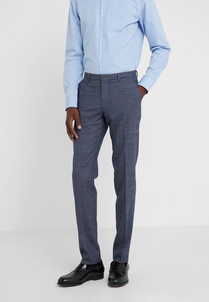 Man som bär blå rutig slim-fit byxor, ljusblå knäppt skjorta, svarta dress skor, står med en hand i fickan mot en vit bakgrund.