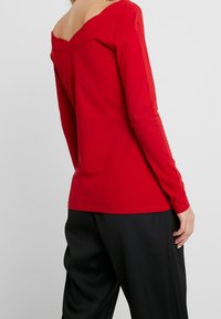 Rotes Langarmoberteil mit weit geschnittenem V-Ausschnitt und glatter Textur, kombiniert mit locker sitzenden schwarzen Hosen. Schlichtes, enganliegendes Design ohne Muster.