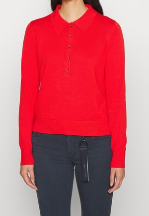 Mujer con un suéter rojo de manga larga y cuello con cinco botones combinado con pantalones oscuros que tienen un llavero negro colgando de un lazo del cinturón.