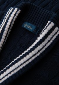 Cardigan lavorato a maglia blu navy con colletto a coste con strisce bianche, texture a trecce e un'etichetta con la scritta "Ombre Heritage" in blu.