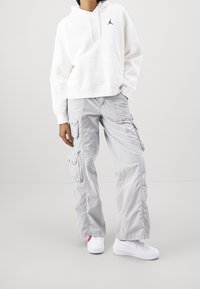 Sudadera blanca con capucha y un logo negro, combinada con pantalones cargo gris claro que tienen varios bolsillos y zapatillas bajas blancas.