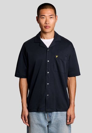 Jeune homme aux cheveux courts portant une chemise noire à manches courtes avec une poche poitrine et un jean bleu clair, debout devant un fond uni.