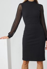 Svart klänning med en åtsittande bodice och knälång kjol. Genomskinliga svarta prickiga ärmar och hög krage. Slät textur och skräddarsydd passform.