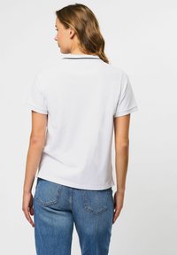 Žena so svetlo hnedými vlasmi v bielom krátkym rukávom polokošeli a modrých džínsach, otočená chrbtom na čistom bielom pozadí.