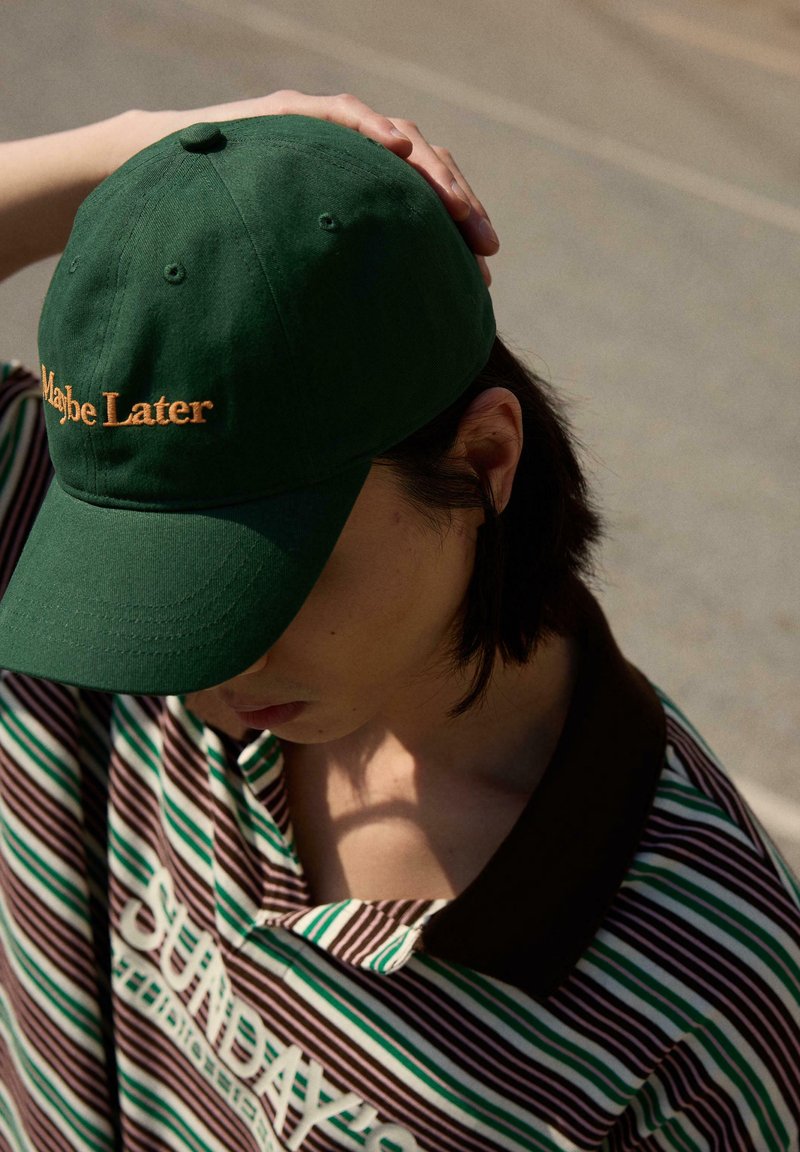 Personne portant une casquette verte avec le texte "Maybe Later" et une chemise rayée avec le mot "SUNDAY" partiellement visible, tête inclinée vers le bas, main sur la casquette.