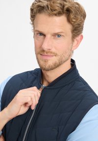 Marineblauwe gewatteerde vest met hoge kraag, voorzien van een rits met leren accent, gedragen over een lichtblauw shirt. Minimalistische ontwerpelementen.