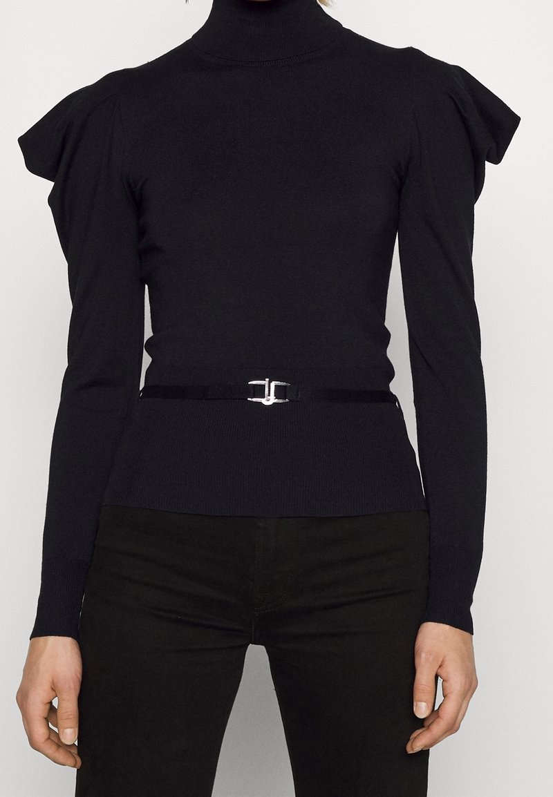 Pull noir ajusté à col roulé avec épaules bouffantes et une fine ceinture noire avec boucle argentée à la taille, porté avec un pantalon noir.