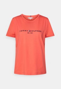 Camiseta color coral hecha de algodón, con un cuello redondo y mangas cortas. En la parte frontal está el texto "TOMMY HILFIGER EST. 1985" en letras azul oscuro.
