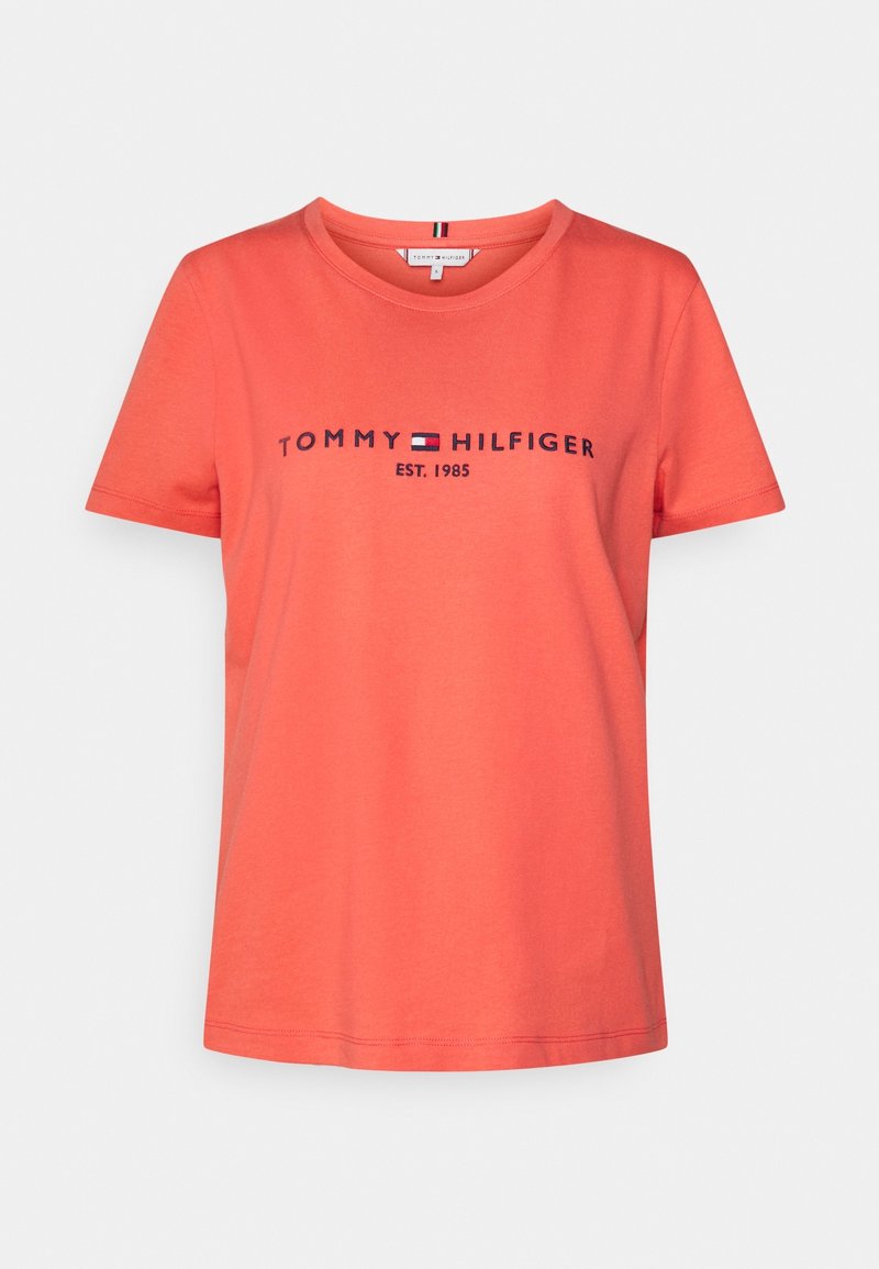 Camiseta color coral hecha de algodón, con un cuello redondo y mangas cortas. En la parte frontal está el texto "TOMMY HILFIGER EST. 1985" en letras azul oscuro.