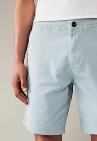 Shorts albastre deschis, dintr-un material moale, cu talie elastică, închidere cu nasture în față și buzunare laterale. Textură netedă și croială ajustată.